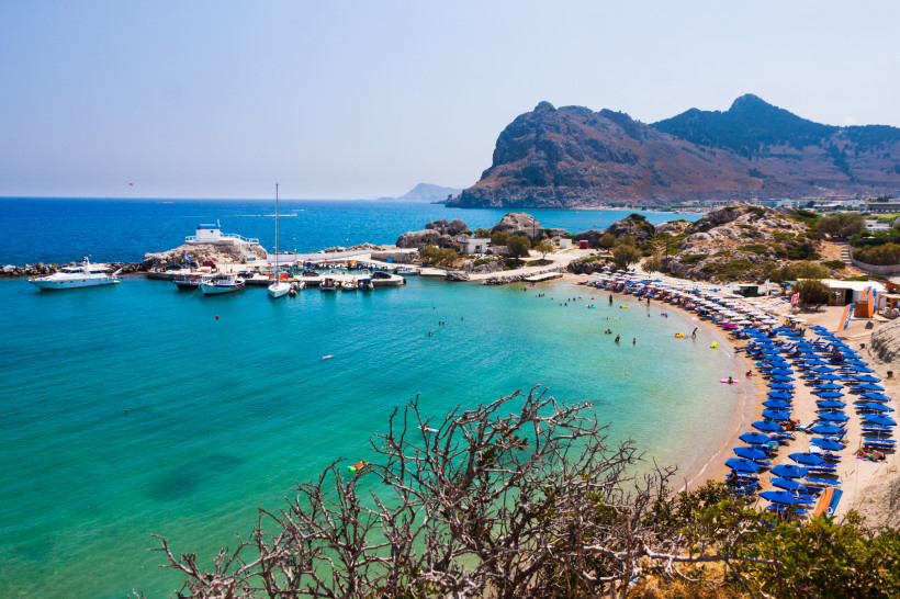 Blick auf eine geschützte Bucht auf Rhodos mit Yachthafen, türkisblauem Meer, Sandstrand mit blauen Sonnenschirmen und felsiger Berglandschaft im Hintergrund
