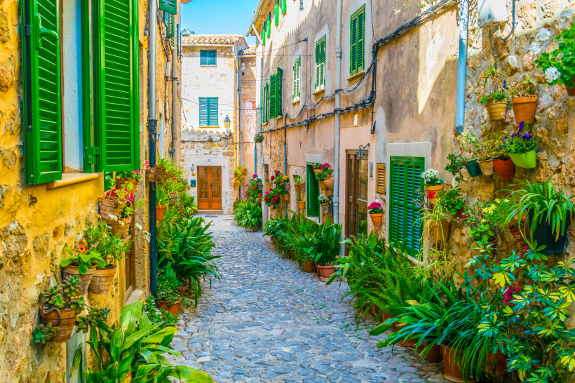 Mallorca - Valldemossa Eine malerische, enge Gasse in einem mediterranen Dorf auf Mallorca. Die Häuser sind aus hellem Stein gebaut und haben grüne Fensterläden. Viele Blumentöpfe und Pflanzen schmücken die Gasse. Die Gasse ist mit Kopfsteinpflaster belegt, was dem Ort eine cha