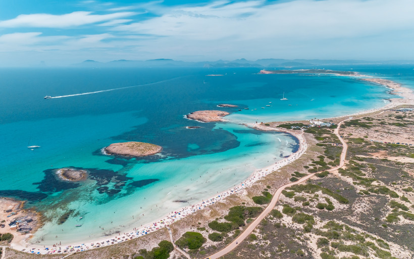 Formentera Luftaufnahme eines weit geschwungenen, weißen Sandstrandes auf Formentera mit türkisblauem Wasser und kleinen vorgelagerten Felsen. Zahlreiche Sonnenschirme säumen den Strand, im Hintergrund ziehen Boote durch das klare Meer. Die Küste ist von Dünen und n