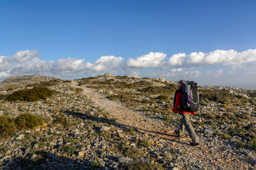 Mallorca - Cami de s´Escolta Das Bild zeigt einen Wanderweg in einer felsigen Berglandschaft bei strahlend blauem Himmel. Im Vordergrund ist ein einzelner Wanderer mit roter Jacke, grauer Hose und einem großen schwarzen Rucksack zu sehen. Er geht auf einem schmalen, steinigen Pfad, d