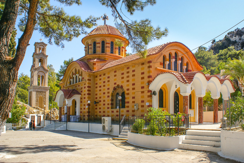 Orthodoxe Kirche auf Rhodos mit rotem Ziegeldach und Glockenturm, umgeben von Pinien und mediterraner Landschaft unter blauem Himmel.