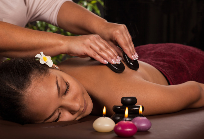 Singapur Eine Frau mit geschlossenen Augen liegt entspannt auf einer Massageliege. Sie erhält eine Hot-Stone-Massage auf dem Rücken. Neben ihr brennen kleine farbige Kerzen, im Vordergrund sind dekorative schwarze Steine gestapelt. Die Frau hat eine weiße Blüte im