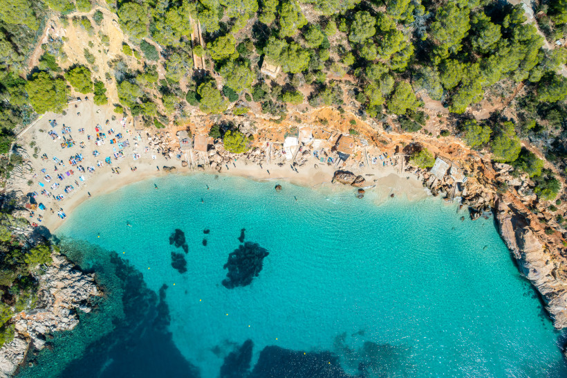 Ibiza - Cala Salada Luftaufnahme der Badebucht Cala Salada auf Ibiza mit türkisblauem Wasser, Sandstrand und umgebenden Pinienwäldern. Am Strand liegen viele Urlauber auf Handtüchern und Liegen.