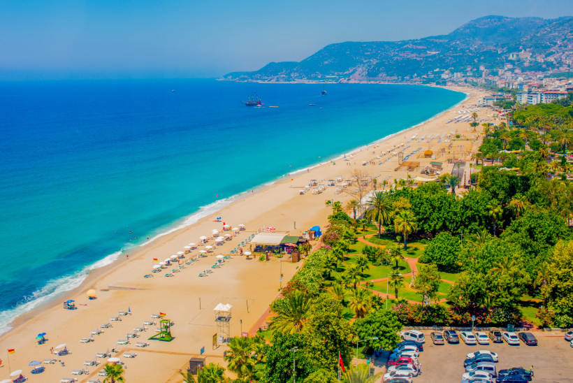 Das Bild zeigt den breiten Cleopatra Strand in Alanya mit klarer Wasserlinie entlang der Küste. Im Vordergrund liegt ein gepflegter Stadtpark mit Wegen und Palmen. Rechts sind Wohnhäuser und Hotelanlagen zu erkennen, dahinter erheben sich bewaldete Berge.