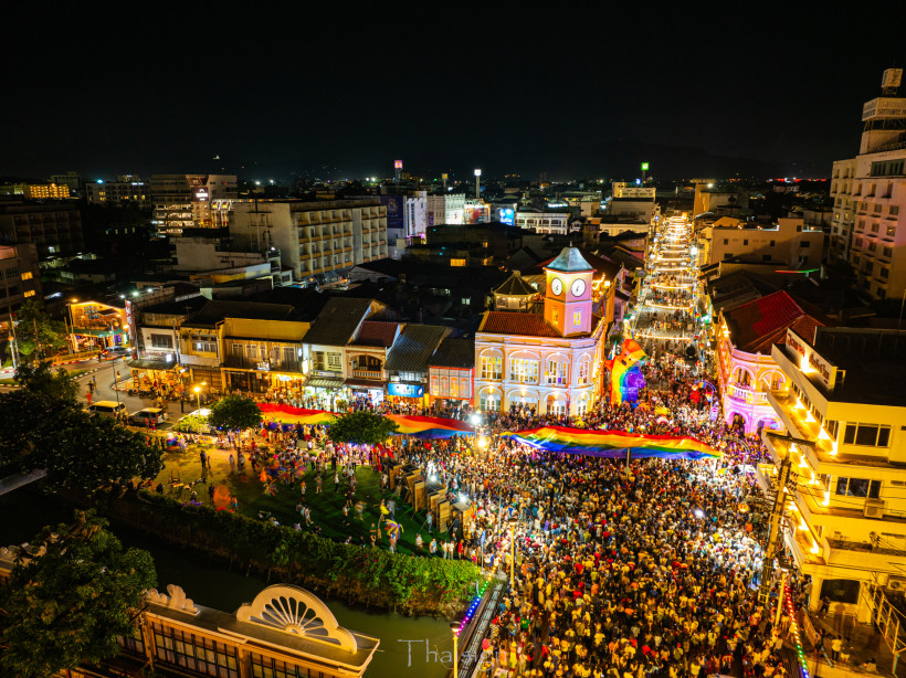 Thailand - Phuket Luftaufnahme des Phuket Pride Festivals bei Nacht. Eine riesige Menschenmenge füllt die geschmückte Straße rund um den ikonischen Uhrenturm. Über dem Festival spannt sich eine große Regenbogenfahne. Die Straße ist hell beleuchtet, Musik und Lichter schaff