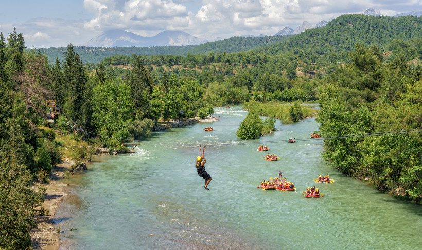 Türkei Person fährt an einer Zipline über einen Fluss, während darunter Gruppen in Schlauchbooten Rafting betreiben, umgeben von grüner Landschaft und Bergen
