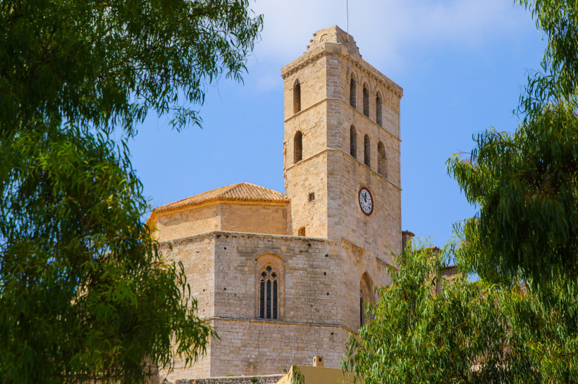 Ibiza Eine historische Kirche aus Sandstein mit einem rechteckigen Turm, in dem mehrere hohe Rundbogenfenster eingelassen sind. Eine große Uhr ist mittig am Turm angebracht. Das Dach ist mit rötlichen Ziegeln gedeckt, und im Vordergrund rahmen grüne Bäume die S
