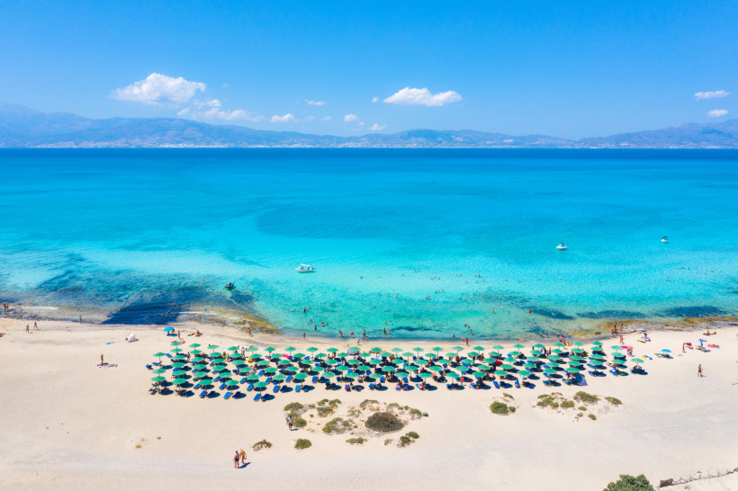Luftaufnahme eines Strandes auf der Insel Chrissi mit türkisfarbenem Meer, Badegästen und Sonnenschirmen
