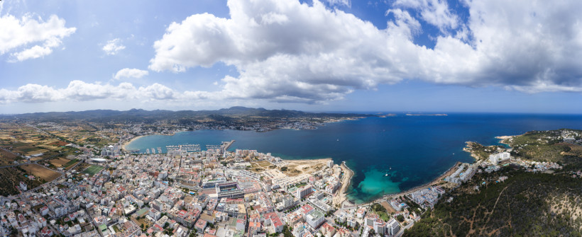 Sant Antoni de Portmany Luftaufnahme von Sant Antoni de Portmany auf Ibiza: Stadt am Meer mit großer Bucht, Marina und Mole; rechts türkisfarbene Buchten, im Hinterland Felder und grüne Hügel unter teils bewölktem Himmel.