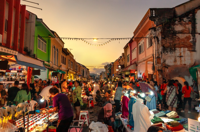 Thailand - Phuket Menschenmengen schlendern über einen lebhaften Nachtmarkt in Phuket Old Town bei Sonnenuntergang. Bunte Kolonialgebäude säumen die Straße, Verkaufsstände bieten Kleidung, Souvenirs und Streetfood an. Warme Lichter hängen über der Straße und sorgen für sti