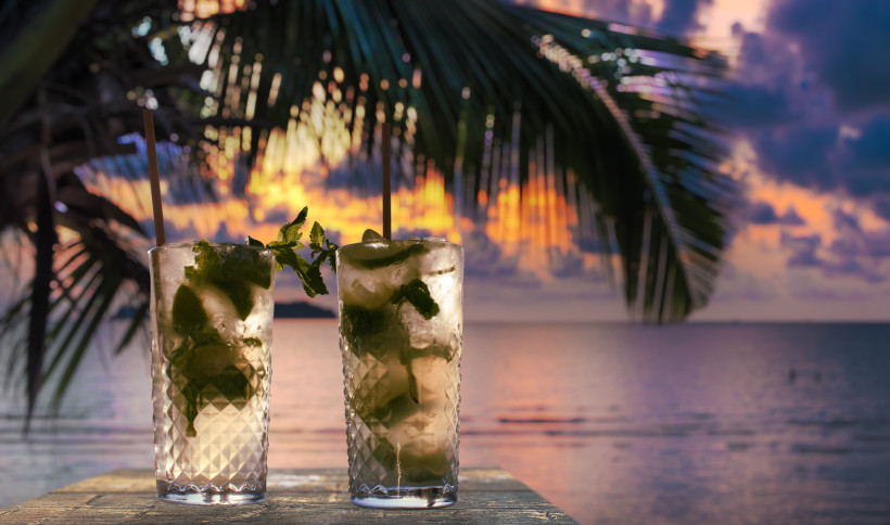 Jamaika Zwei Mojito-ähnliche Cocktails mit Limetten, Minze und Eis stehen auf einem Holztisch am Meer. Im Hintergrund geht die Sonne über dem Wasser unter und taucht den Himmel in dramatische Farben aus Violett, Orange und Gold. Eine große Palmenwedel ragt von li