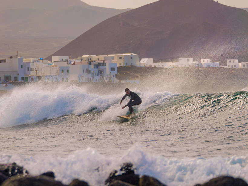 Surfer in La Santa – Beliebter Surfspot an Lanzarotes Westküste Surfer reitet eine Welle vor La Santa auf Lanzarote, im Hintergrund weiße Häuser und vulkanische Berge.