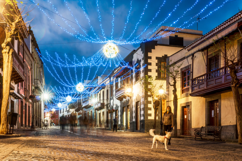 Straße in Teror auf Gran Canaria am Abend, festlich geschmückt mit leuchtenden Lichterketten, Menschen spazieren entlang der traditionellen Häuserfassaden, im Vordergrund ein Hund.