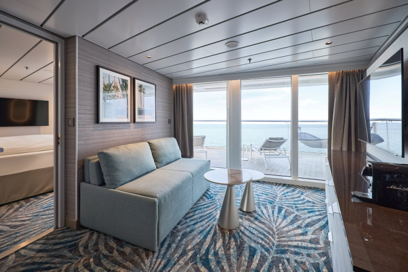 Mein Schiff Relax – Fernweh Suite: Wohnbereich mit Glasfront zur Terrasse und Meerblick Wohnbereich der Fernweh Suite auf Mein Schiff Relax mit Sofa, Couchtisch und Blick durch die Glasfront auf die Terrasse und das Meer