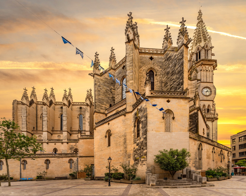 Mallorca - Manacor Eine beeindruckende Kirche im neugotischen Stil, wahrscheinlich in Spanien oder Südfrankreich, mit einer Fassade aus hellem Sandstein. Die Kirche hat viele spitze Türme und Zinnen, die an eine Burg erinnern. Blaue und weiße Fahnen sind an einem Seil zwisc