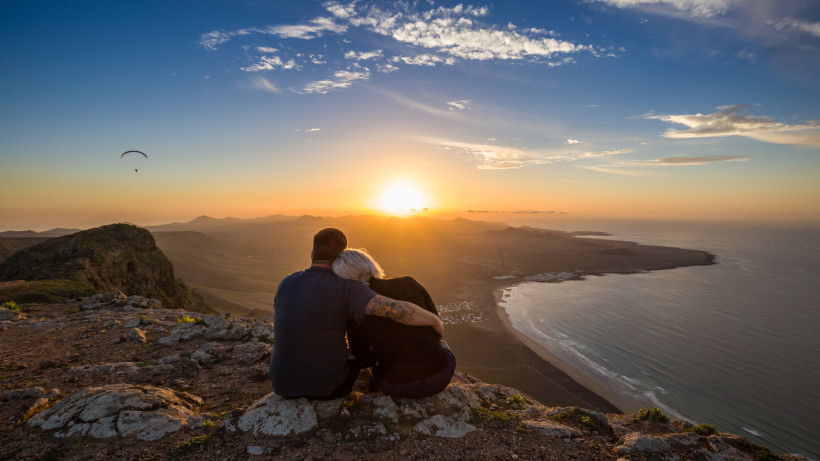 Sonnenuntergang auf Lanzarote – Romantischer Aussichtspunkt für Paare Paar sitzt auf einer Klippe und genießt den Sonnenuntergang über der Küste von Lanzarote – romantischer Moment für Paare und Erwachsene auf der Kanareninsel.