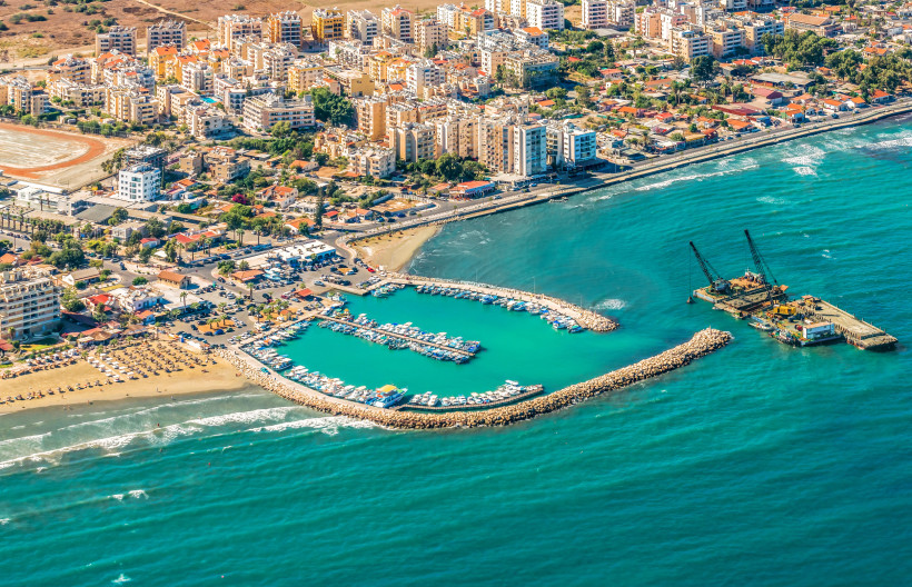 Larnaca Marina aus der Luft: halbkreisförmiges Hafenbecken mit Yachten, Strandabschnitt und Stadtpanorama an Zyperns Südküste.