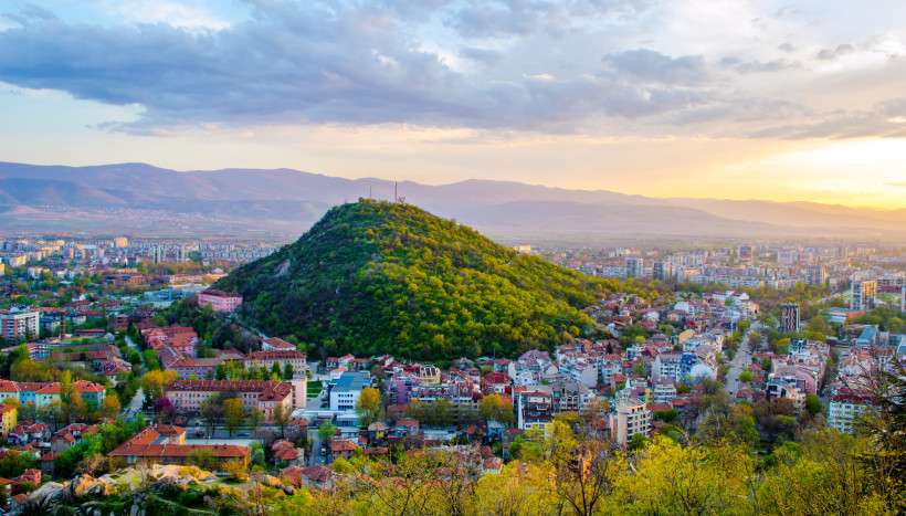 Panoramablick auf Plovdiv bei Sonnenuntergang. Im Zentrum ein bewaldeter Hügel mit Antennenanlagen, umgeben von Wohnhäusern, Bäumen und modernen Stadtteilen. Im Hintergrund liegen Berge unter einem farbigen Abendhimmel.