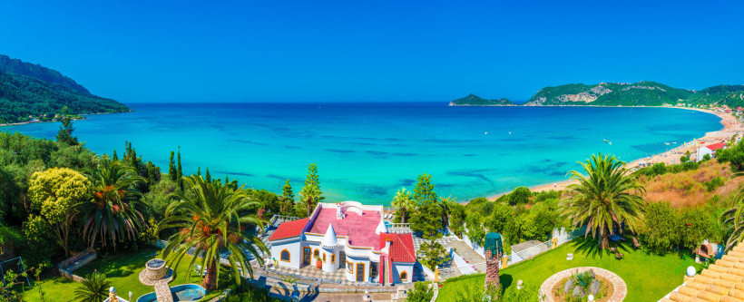 Küstenlandschaft auf Korfu mit türkisblauem Meer, Sandstrand und weißem Haus mit rotem Dach im Vordergrund