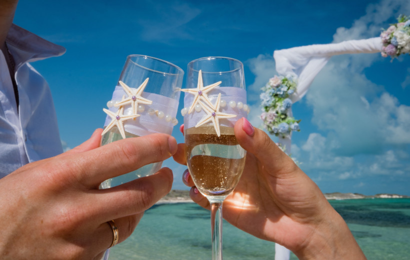 Hände eines Paares stoßen mit dekorierten Sektgläsern am Strand an, Meerblick im Hintergrund