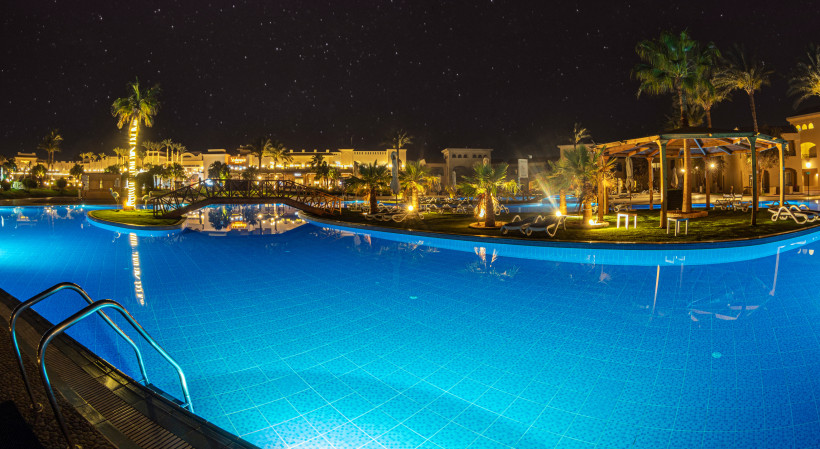 Stimmungsvoll beleuchtete Poollandschaft eines Resorts unter dem Sternenhimmel Beleuchteter Hotelpool bei Nacht mit Palmen, Holzbrücke und klarer Spiegelung im Wasser