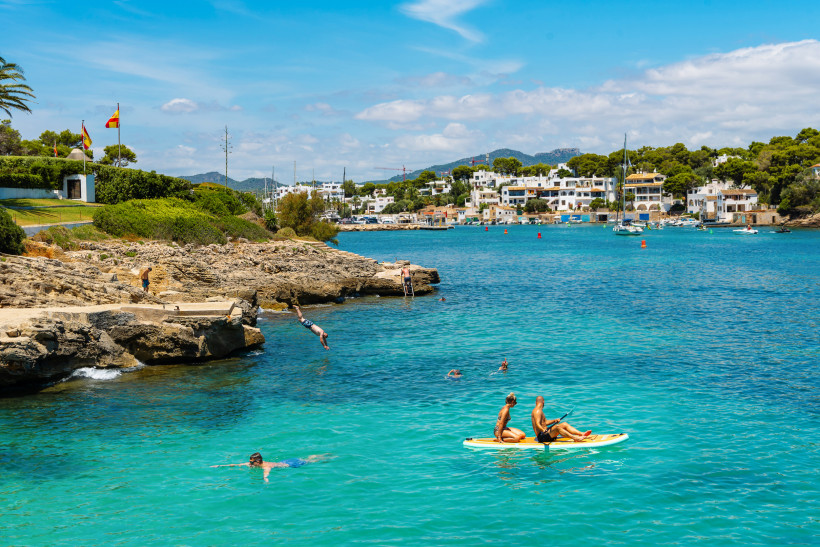 Cala d’Or, Mallorca Badegäste im türkisblauen Wasser von Cala d’Or auf Mallorca, mit Felsen, Klippenspringern und Stand-Up-Paddlern vor weißen Häusern am Ufer