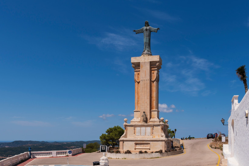 Christusstatue mit ausgebreiteten Armen auf einem hohen Steinsockel auf dem Monte Toro, dem höchsten Punkt Menorcas. Die Statue steht auf einem Aussichtsplateau mit Blick über die Insel. Der Himmel ist klar blau, es sind nur wenige kleine Wolken zu sehen.