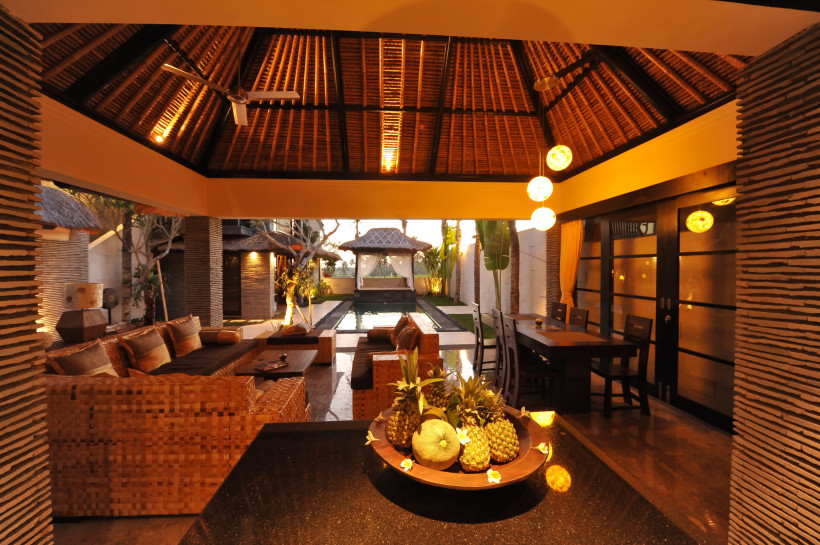 Thailand - Koh Samui Das Bild zeigt den Innenbereich einer offenen, luxuriösen Villa im tropischen Stil. Das hohe Dach ist mit traditionellen Naturmaterialien gedeckt. Im Vordergrund steht eine schwarze Steinplatte mit einer dekorativen Obstschale, gefüllt mit Ananas, Melone