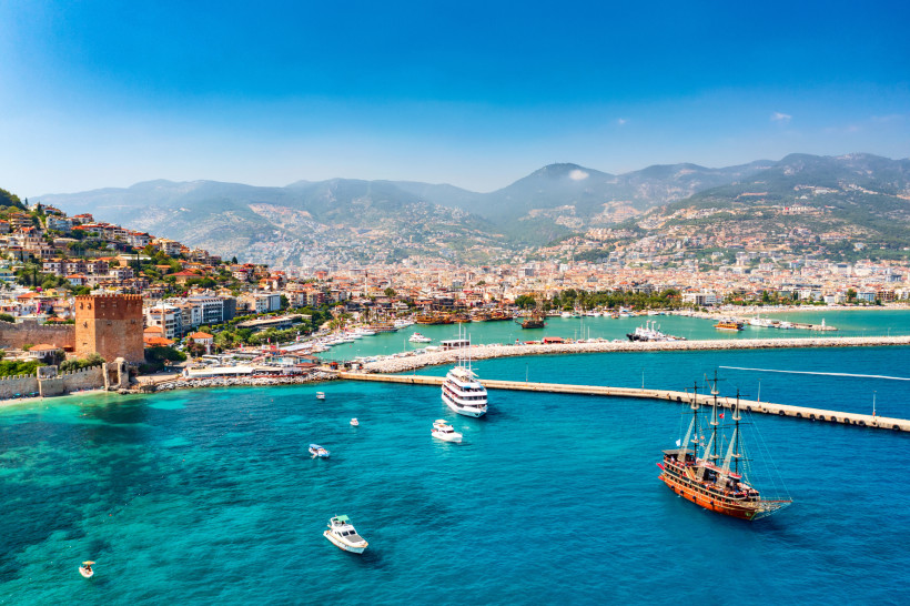 Auf dem Bild ist der Hafen von Alanya zu sehen, mit dem markanten Roten Turm im Vordergrund. Im Wasser liegen Segelschiffe und Yachten. Die Stadt breitet sich dahinter am Hang aus, mit bewaldeten Bergen im Hintergrund.