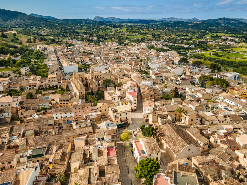 Mallorca - Son Servera Luftaufnahme von Son Servera mit dem historischen Zentrum, der Església Nova und den umliegenden Wohnhäusern. Die Häuser haben meist sandfarbene Fassaden und Terrassen. Im Hintergrund weite Felder, grüne Hügel und Berge bis zum Horizont. Eine ruhige, länd