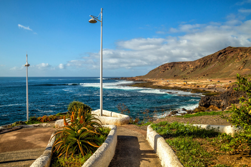 Küstenlandschaft am El Confital mit Promenade, Laternenmast, Pflanzenbeet und Brandung vor felsiger Küste
