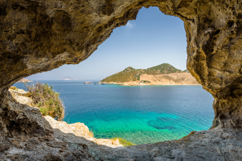 Kos Blick durch eine natürliche Felsenöffnung auf eine türkisfarbene Bucht bei Kos. Im Hintergrund liegt eine kleine, hügelige Insel mit grün-brauner Vegetation, an deren Küste zwei Boote ankern. Das Wasser ist klar und schimmert in verschiedenen Blautönen.