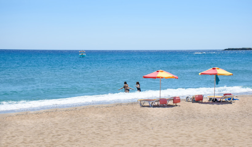 Sandstrand auf Rhodos mit zwei badenden Personen im Meer, bunten Sonnenschirmen, Liegen und einem Boot auf dem offenen Wasser
