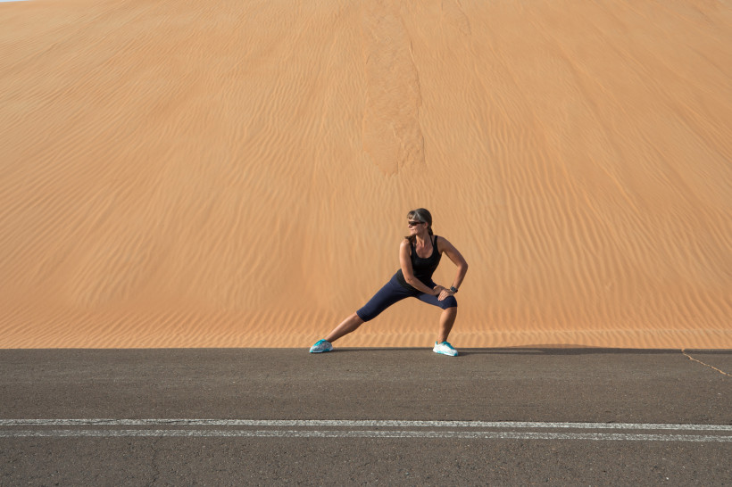 Frau in Sportkleidung dehnt sich auf einer Straße vor einer riesigen Sanddüne in der Wüste.