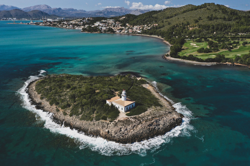 Luftaufnahme der Halbinsel Alcanada mit Leuchtturm, felsiger Küste und Blick über die Bucht von Alcudia bis zur Bebauung von Port d'Alcudia
