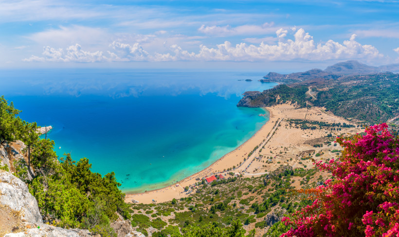 Das Bild zeigt eine weite Panoramansicht auf eine atemberaubende Küstenlandschaft auf einer griechischen Insel – wahrscheinlich Kreta oder Rhodos.    Im Vordergrund wächst eine leuchtend pinke Bougainvillea. Rechts unten beginnt ein goldgelber Sandstrand,