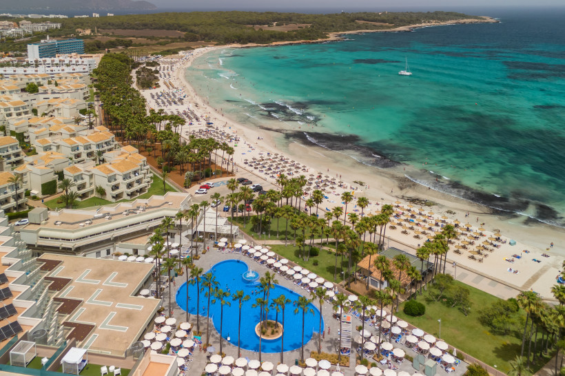Luftaufnahme von Sa Coma mit Sandstrand, Sonnenschirmen, türkisfarbenem Meer und Hotelanlage mit Pool