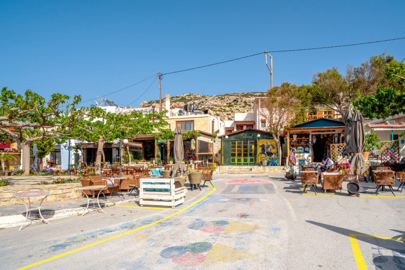 Kreta Ein kleiner, bunter Platz im Ort Matala auf Kreta mit Tavernen, Cafés und kleinen Läden. Im Vordergrund stehen Tische und Stühle unter schattigen Bäumen, im Hintergrund bunte Hausfassaden und ein Souvenirladen. Der Himmel ist klar und blau, das Licht warm