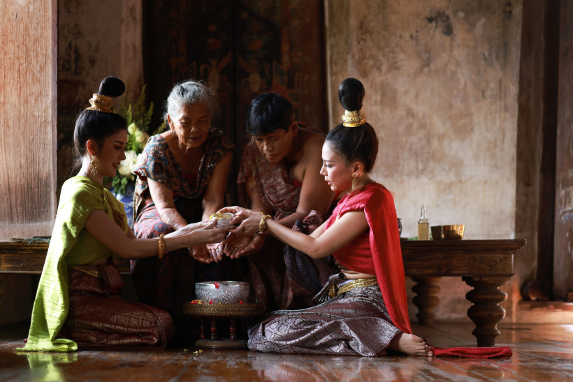 Zwei junge Frauen in traditioneller thailändischer Kleidung gießen Wasser über die Hände zweier älterer Menschen. Alle sitzen respektvoll am Boden in einem historischen Raum. Eine silberne Schale mit Blüten steht vor ihnen.
