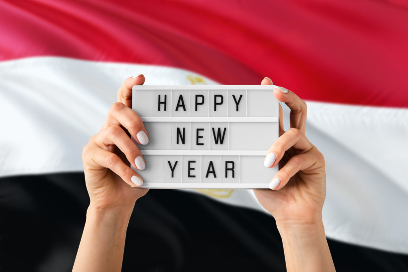 Ägypten Zwei Hände halten ein Leuchtschild mit der Aufschrift 'HAPPY NEW YEAR' vor einer wehenden ägyptischen Flagge in Rot, Weiß und Schwarz.