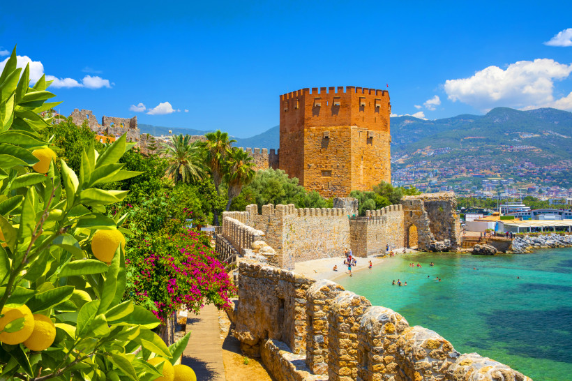 Der Rote Turm (Kızıl Kule) in Alanya, eine mittelalterliche Festung direkt am Meer. Im Vordergrund sind Zitronenbäume, Bougainvillea und Palmen zu sehen. Entlang der alten Stadtmauer baden Menschen im türkisblauen Wasser. Im Hintergrund liegt die Stadt Al