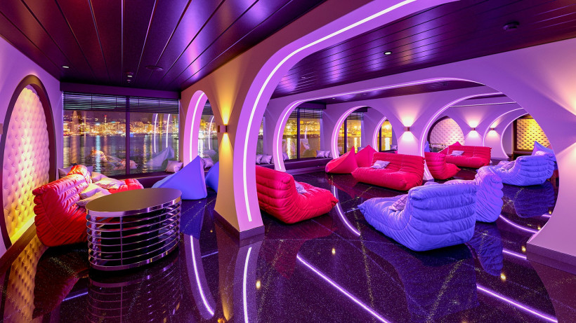 Wave Club Teens Lounge an Bord von AIDAcosma und AIDAnova mit Sitzsäcken, LED-Lichtbögen und Blick auf Hafenlichter Innenraum des Wave Club Teens Lounge auf einem AIDA-Kreuzfahrtschiff mit farbiger LED-Beleuchtung, Sitzsäcken und Panoramafenstern bei Nacht