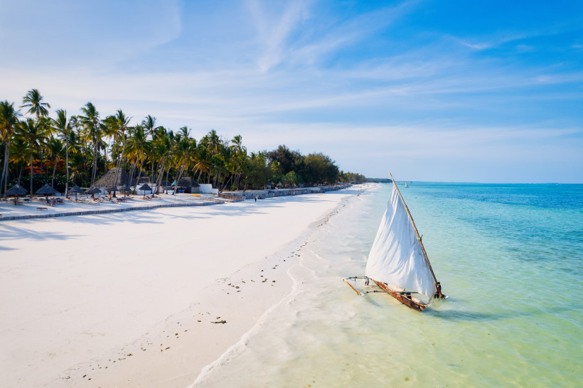 Sandstrand auf Sansibar mit Palmen und Segelboot im flachen, türkisfarbenen Meer