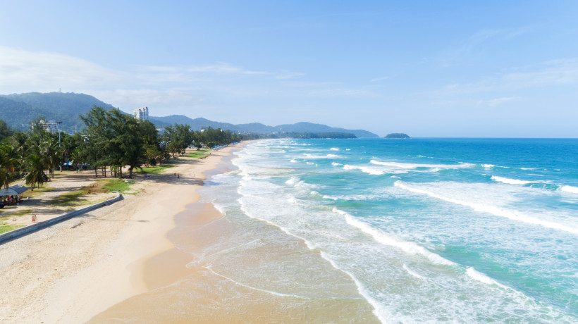 Blick über den langen Sandstrand von Karon Beach mit Brandung, türkisfarbenem Meer und Hügeln im Hintergrund