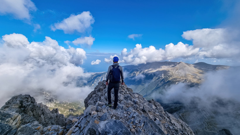 Griechenland - Olymp Bergsteiger mit Helm und Rucksack auf einem Gipfel des Olymps, umgeben von Wolken und Blick auf die umliegenden Berge.