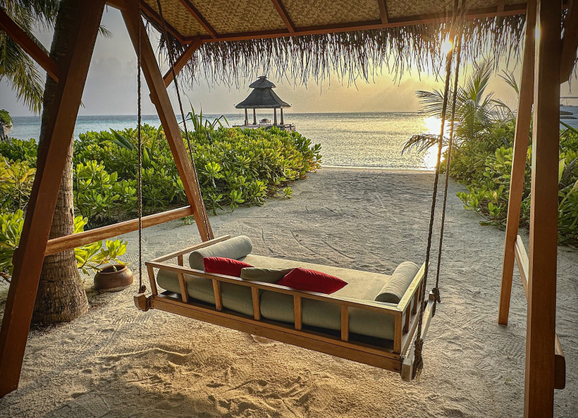 Malediven -  Baros Maldives Romantisches Schaukelbett mit Kissen unter einem Palmendach am Strand der Malediven mit Blick auf den Sonnenuntergang und das Meer.