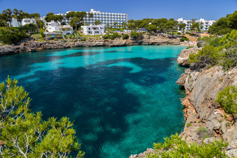 Geschützte Felsbucht in Cala d’Or mit kristallklarem Wasser, Hotelanlage und Badegästen am kleinen Sandstrand