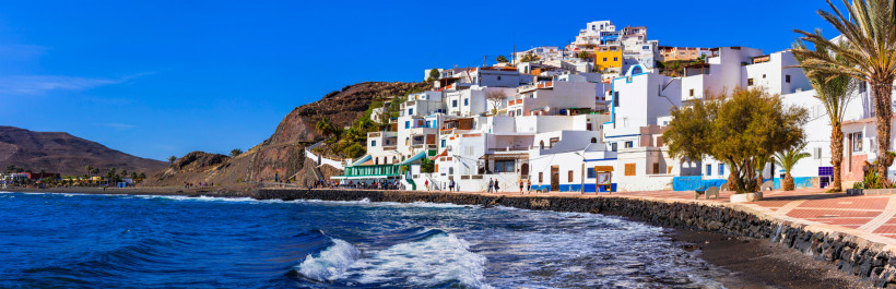 Las Playitas: Weißes Küstendorf am Hang mit Promenade und Atlantikwellen Das charmante Fischerdorf Las Playitas zeigt sich hier mit seinen weiß-blauen Häusern, die sich malerisch an den Hang schmiegen. Die schmale Promenade, das dunkle Vulkangestein und der weite Atlantik formen eine Kulisse, die typisch für den ruhigen Osten