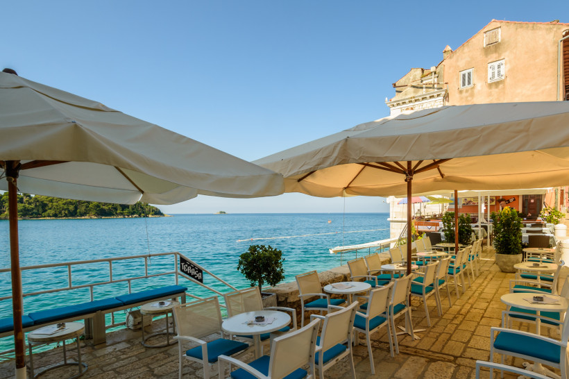 Gemütliche Restaurantterrasse mit weißen Stühlen und Sonnenschirmen direkt am Wasser in Rovinj, Kroatien. Blick auf das türkisfarbene Meer und eine kleine Insel am Horizont.