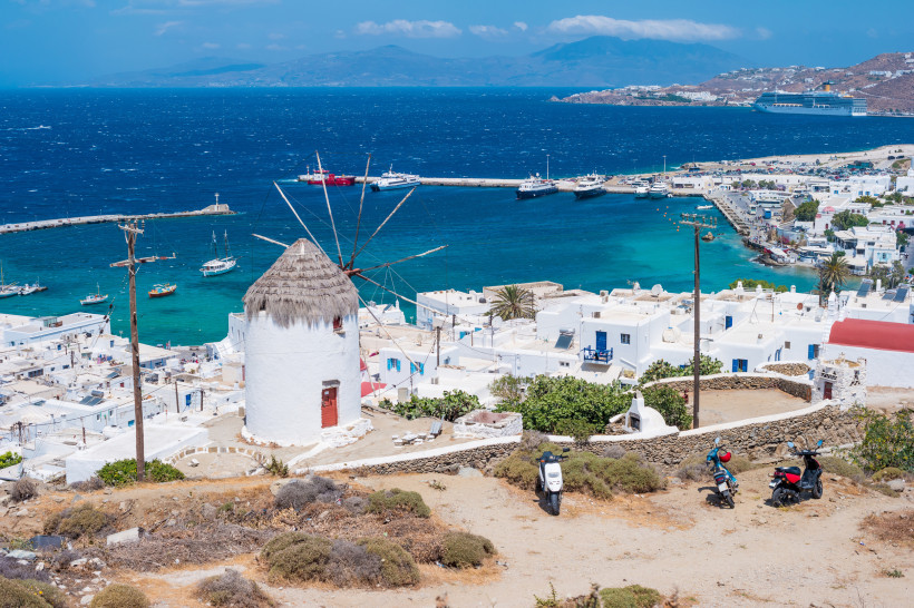 Traditionelle weiße Windmühle mit Strohdach auf einer Anhöhe mit Blick auf eine Stadt mit weißen Häusern und den blauen Ozean. Im Hafen liegen zahlreiche Boote und ein Kreuzfahrtschiff. Der Himmel ist klar und sonnig.